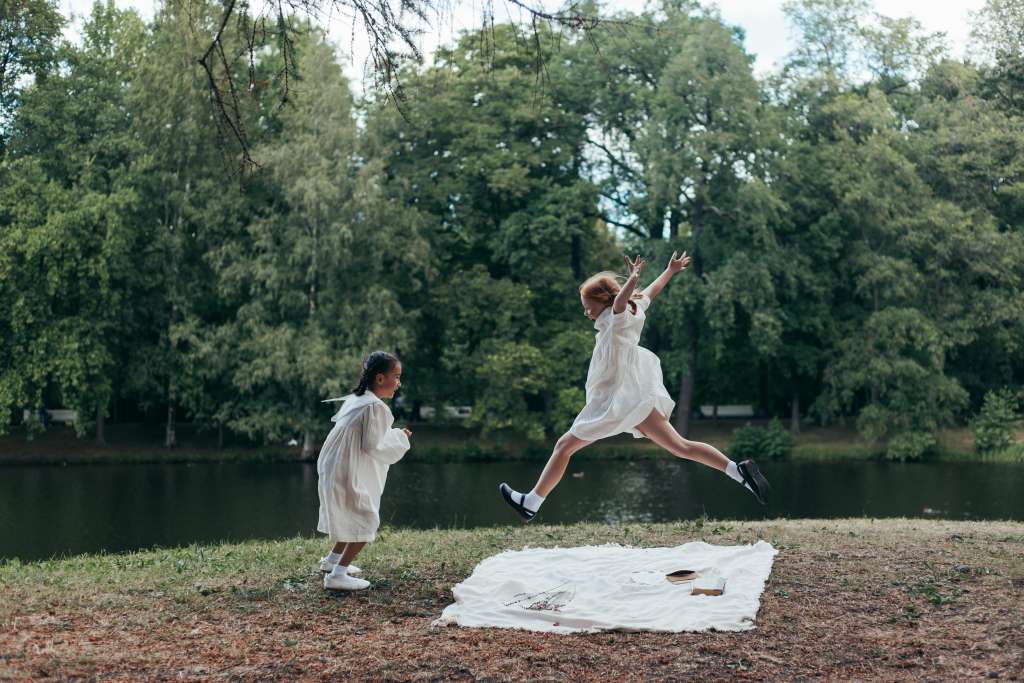Two girls in white dresses playing joyfully by a lake in a lush green park.