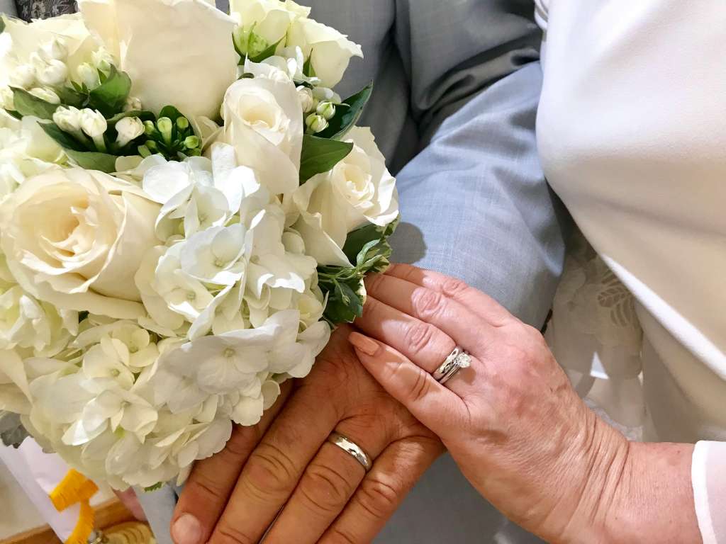 A close-up of newlyweds' hands with rings, holding a white floral bouquet, symbolizing love and commitment.